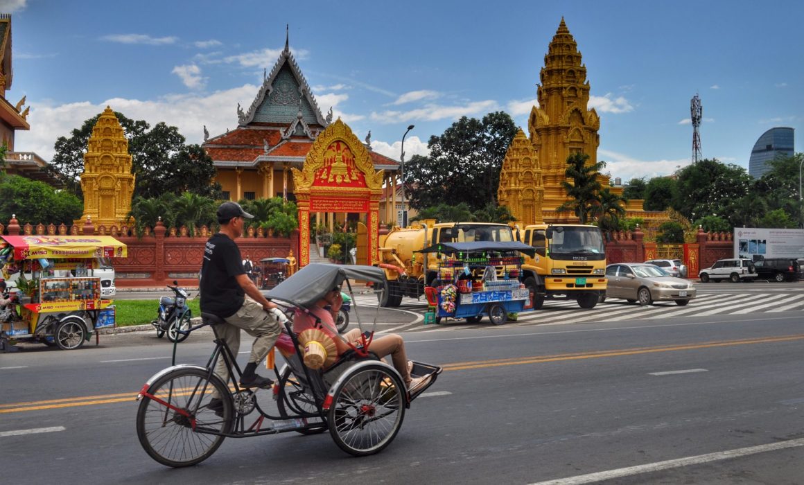 Man in Grasshopper Adventures t shirt pedaling rickshaw past pagoda in Phnom Penh