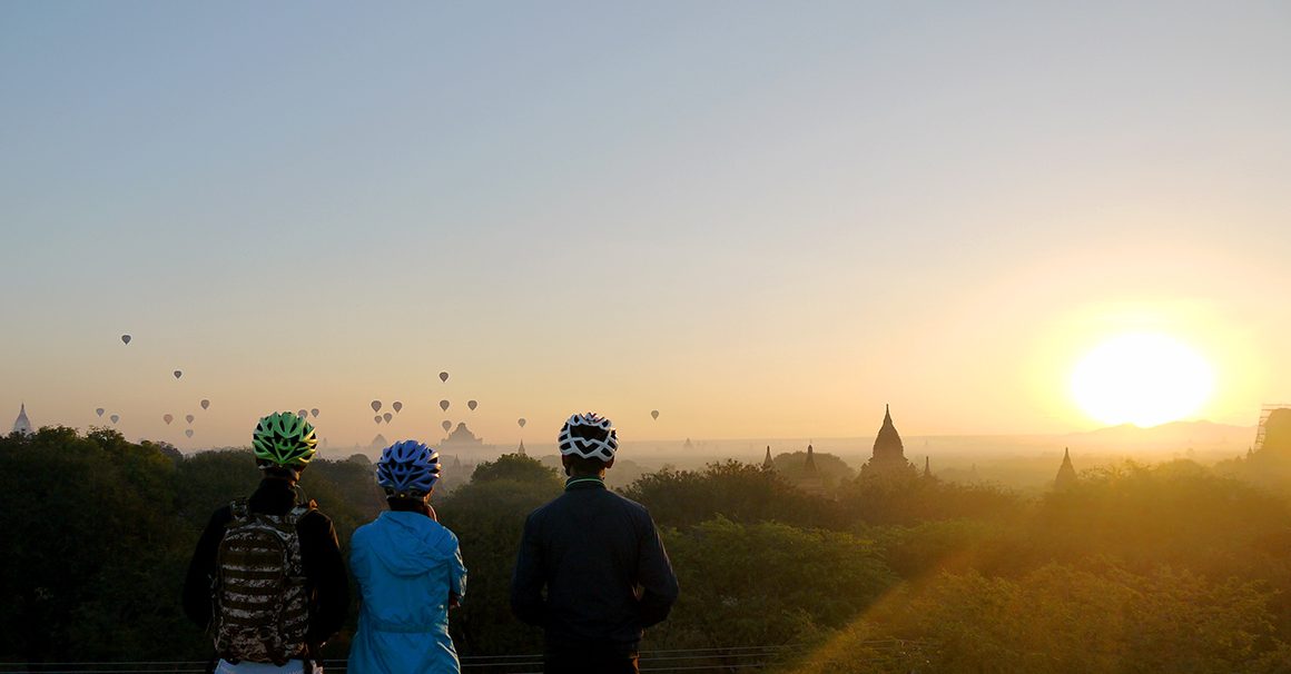 Three cyclists in helmets watching balloons and sunrise over temples of Bagan