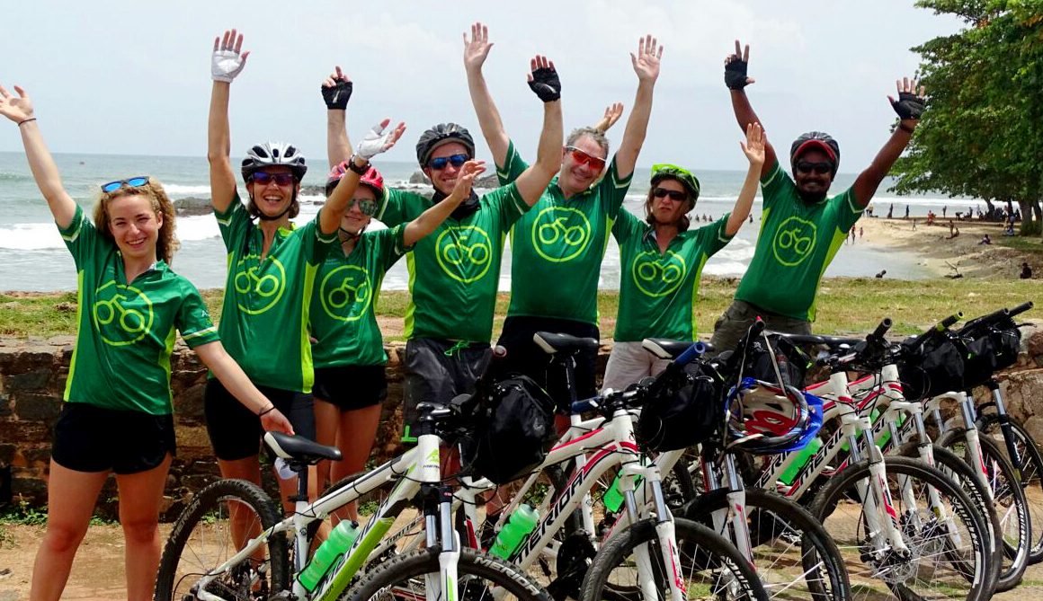 Seven cyclists in Grasshopper Adventures cycling jerseys with raised arms in front of beach