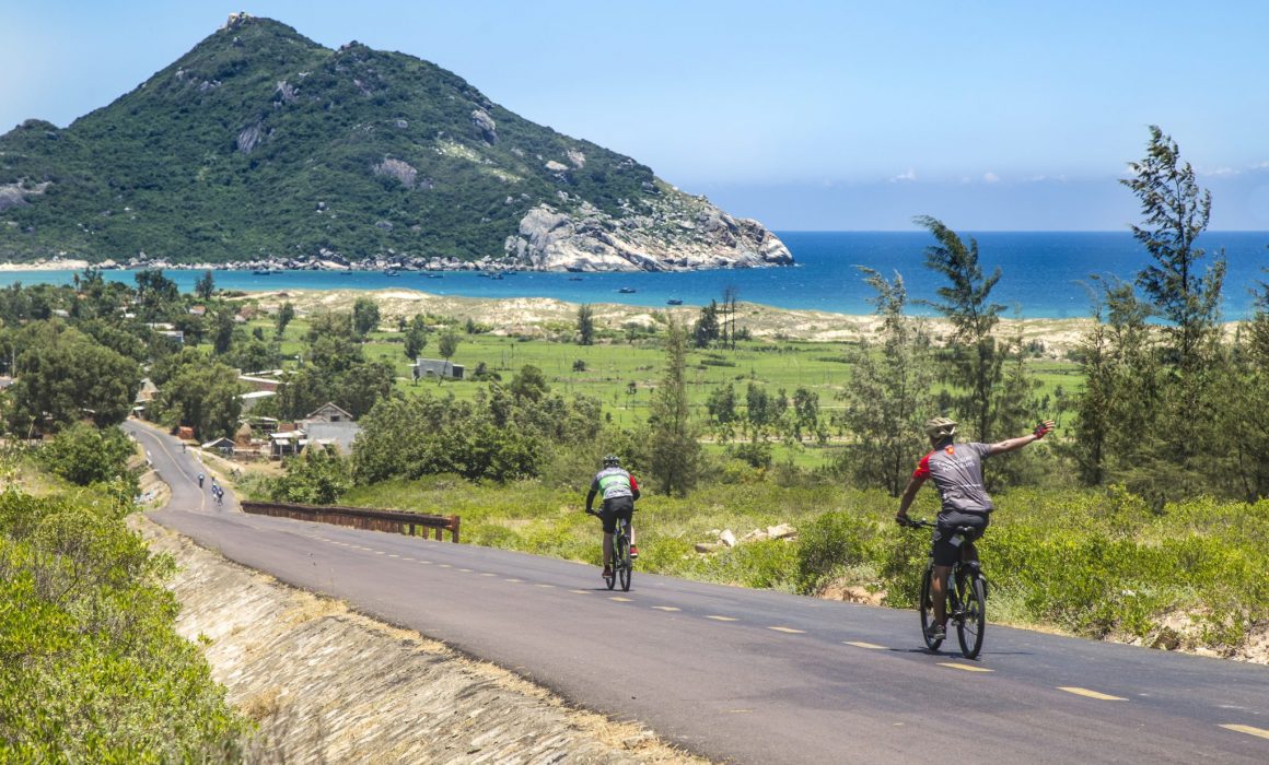 Two cyclists riding on concrete road with bright blue water and hill island in the background