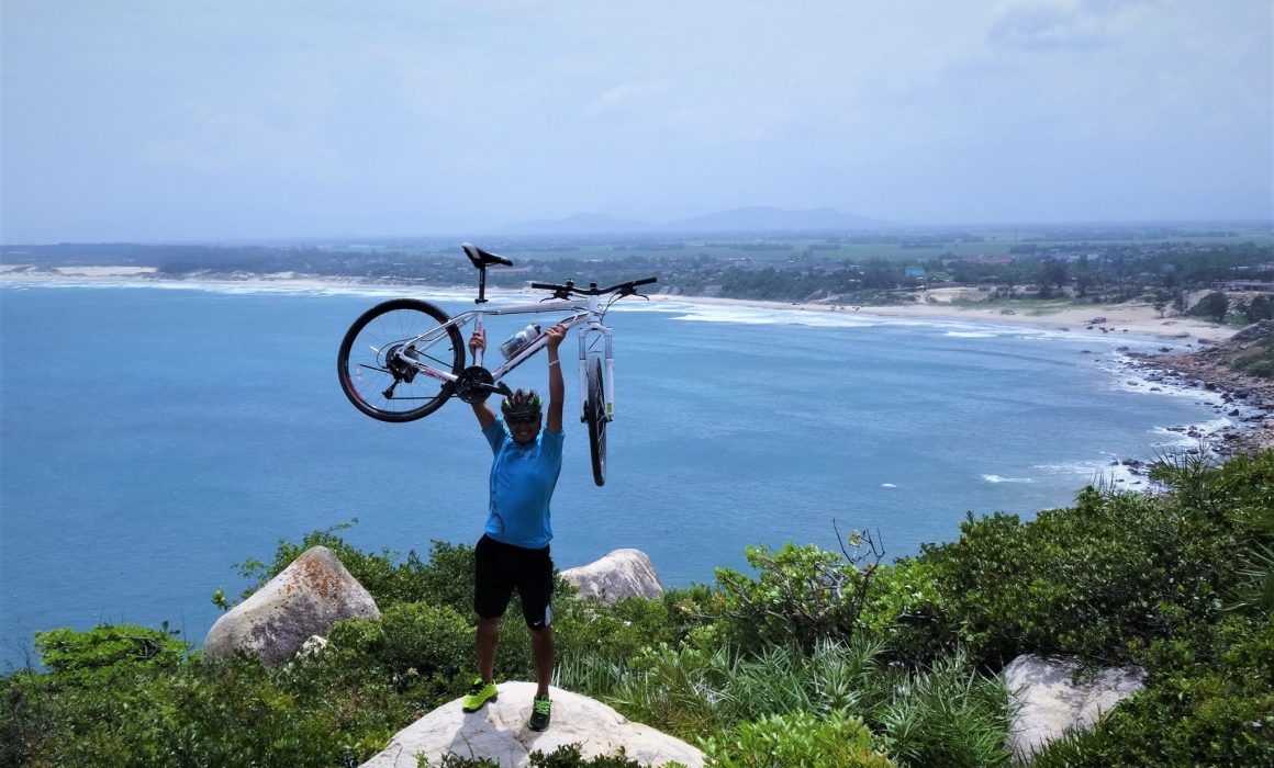 Cyclist raising bike above head with water and coastline in the background