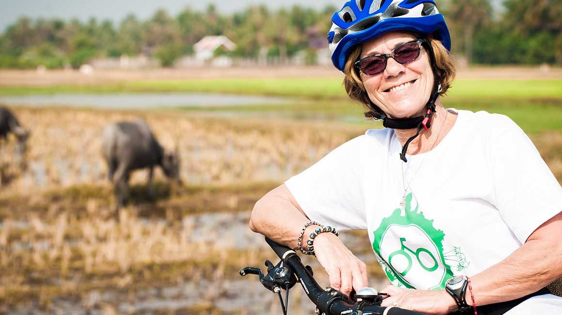 Smiling female cyclist leaning on bike in helmet in front of fields where buffalo are grazing in Siem Reap countryside