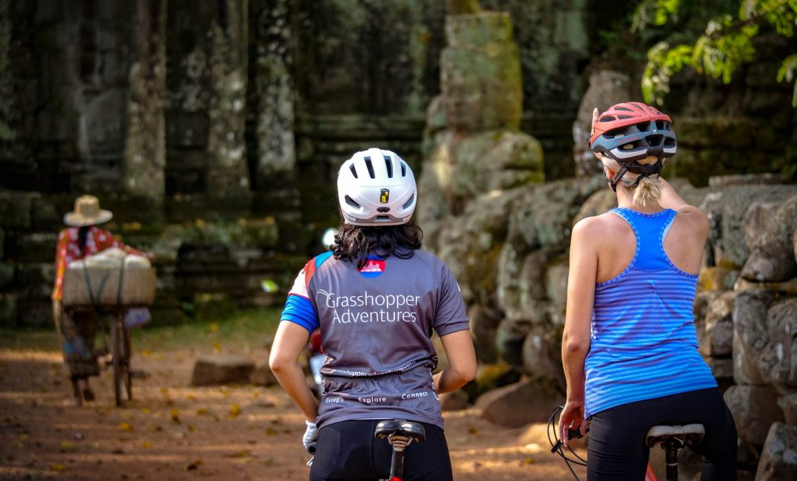 Two cyclists, one in Grasshopper Adventures Cambodia cycling jersey, pointing at temple ruins ahead and local woman on bike ahead