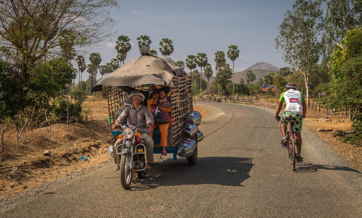 Cyclist in Cambodia cycling jersey riding past cart filled with locals on countryside road of Cambodia