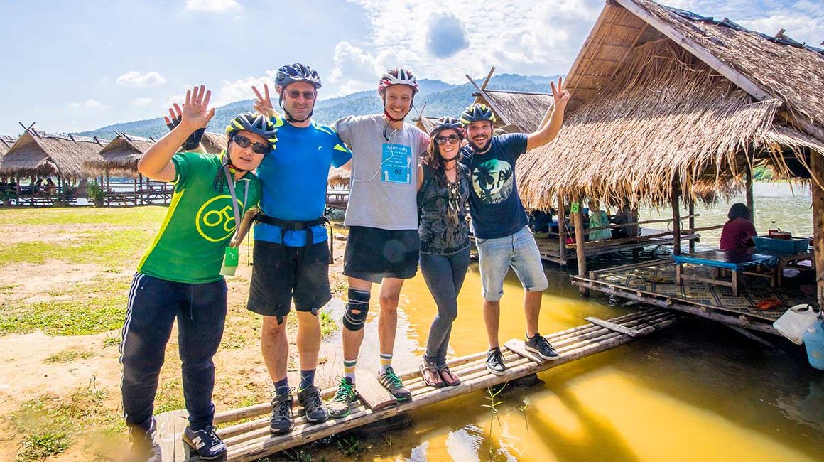 Guide and four cyclists in helmets with hands in the air standing on a walkway to a hut floating on water outside of Chiang Mai