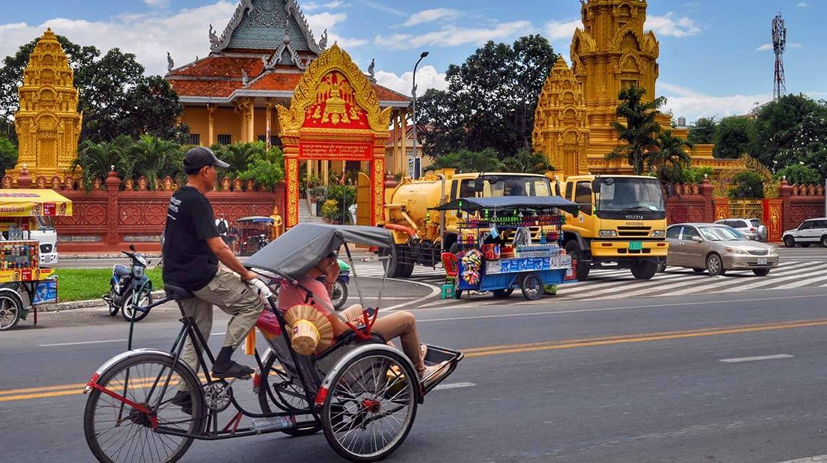 Local man in Grasshopper Adventures t shirt pedaling rickshaw past pagoda on street of Phnom Penh