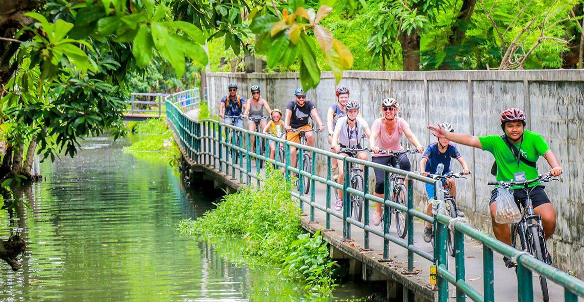 Eight cyclists following tour guide on concrete path by canal in Bangkok