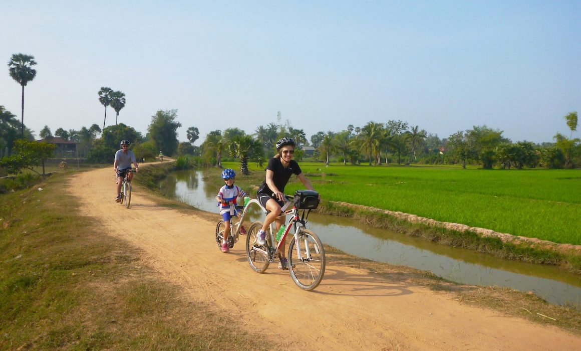 Family with child on tagalong bike on dirt path through countryside of Cambodia