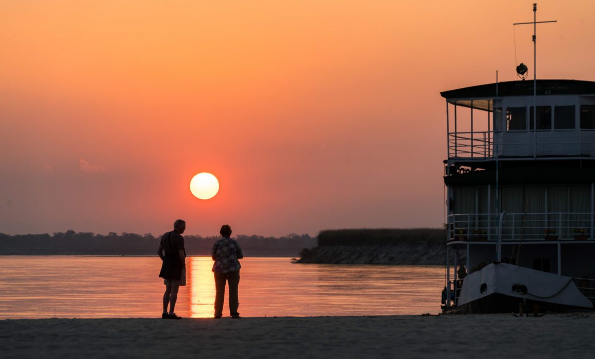 Two people standing under sun and its reflection on the water with boat nearby