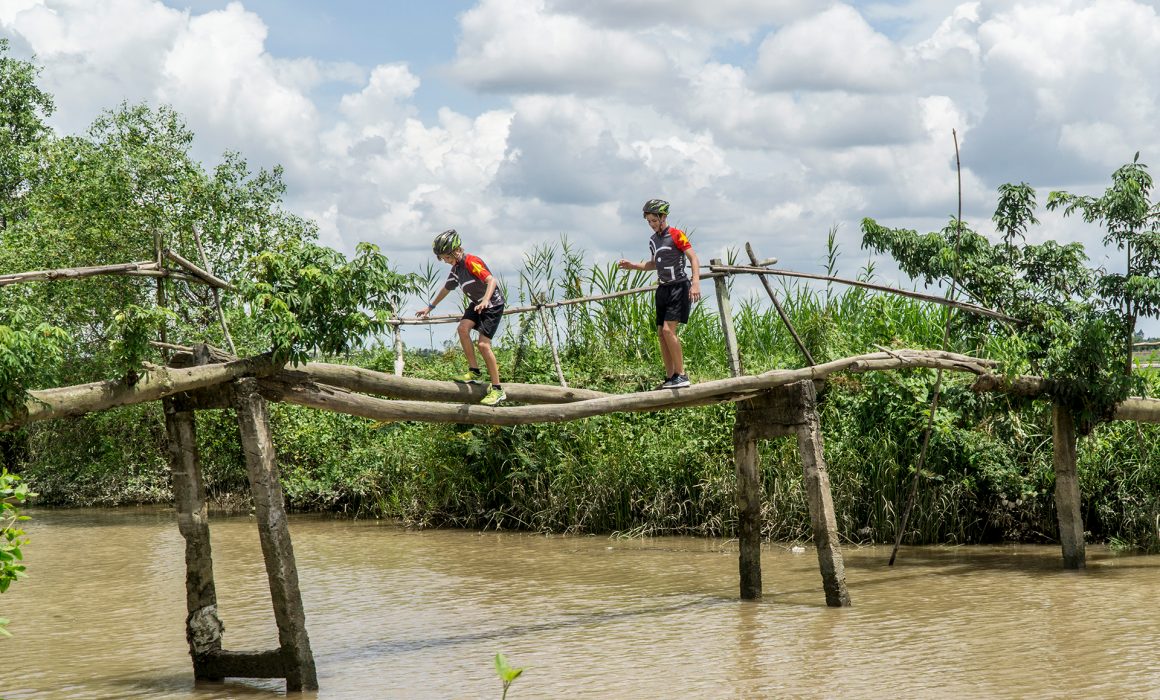 Two people in Grasshopper Adventures Vietnam cycling jerseys and helmets crossing wooden bridge in Vietnam