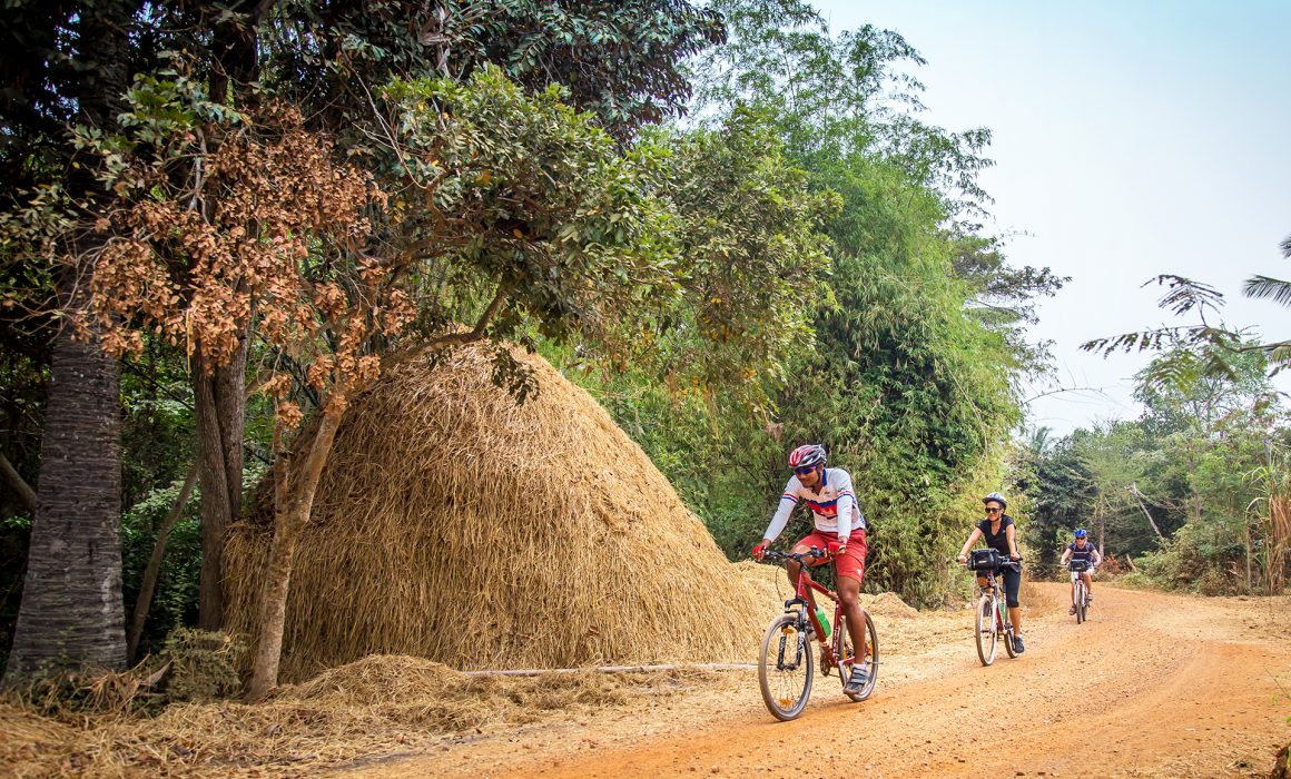 Three cyclists riding on dirt road past trees and haystack in countryside of Cambodia